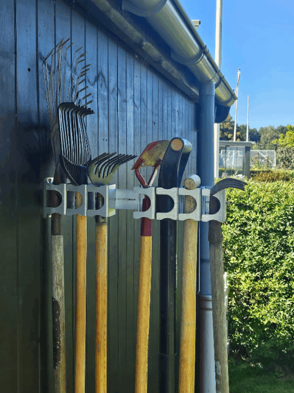 Viting-redskabsophæng-7-4 Et smart haveredskabsophæng i metal på en sort trævæg der holder 7 redskaber med hovedet opad. Ophænget har individuelle holdere til hvert redskab hvor den ene holder er åben.