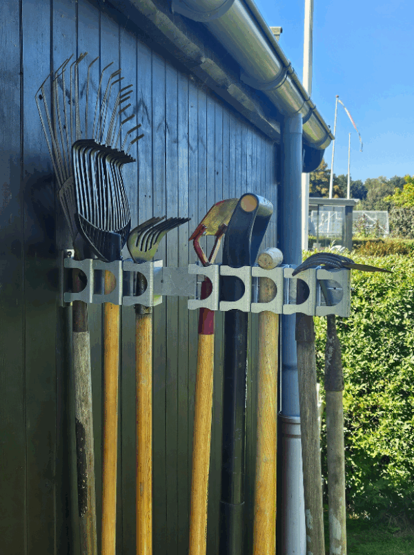 Et haveredskabsophæng til 8 redskaber i metal på en sort trævæg der holder 8 redskaber med hovedet opad. Ophænget har individuelle holdere til hvert redskab hvor den ene holder er åben.